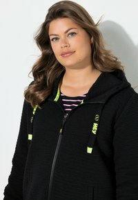 Sudadera con capucha negra con cremallera, de tela texturizada, con cordones y detalles en verde neón. Lleva puesta sobre una camisa de rayas rosas.