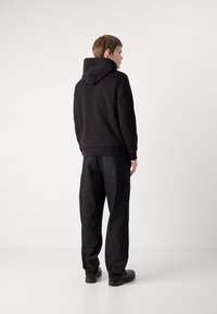 Svart bomullshoodie med snøring i hetten, kombinert med løse svarte denimbukser, sett fra baksiden. Jevn tekstur, minimalistisk design.