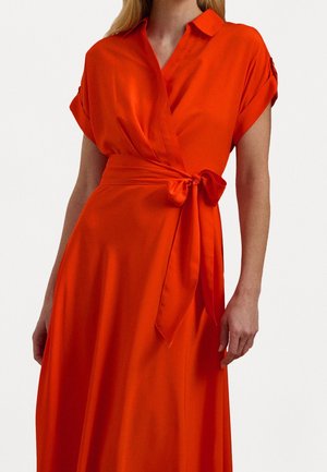 Femme portant une robe portefeuille orange vif avec des manches courtes retroussées et un nœud noué à la taille, sur un fond uni.