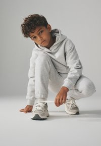 Chłopiec z kręconymi włosami, ubrany w szary hoodie i dresy, klęczy, związując sznurówki w białych trampkach na tle jednolitego tła.