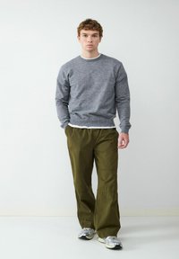 Grauer Strickpullover mit Rundhalsausschnitt, kombiniert mit olivgrünen, weiten Hosen. Das Model trägt graue und schwarze Sneaker. Schlichter Hintergrund.