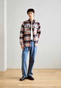Camicia di flanella a quadri arancione, blu e nera, con due tasche sul petto, abbinata a jeans azzurri chiari e scarpe nere slip-on.