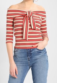 Off-shoulder stribet top i rød og beige, ribbet tekstur, med en frontknude detalje og trekvartærmer. Parret med denim jeans.