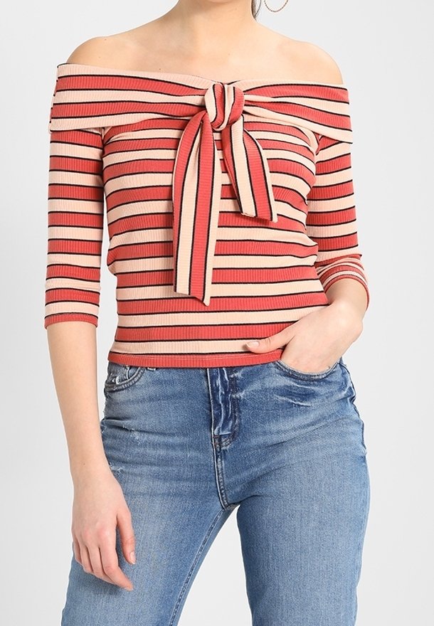 Off-shoulder stribet top i rød og beige, ribbet tekstur, med en frontknude detalje og trekvartærmer. Parret med denim jeans.