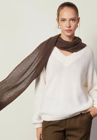 Une écharpe marron en tissu doux drape un pull en maille blanche à col en V. Le modèle porte un pantalon marron, mettant en valeur un look simple et superposé.