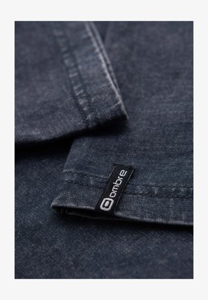 Tissu en denim bleu foncé avec une texture lisse, présentant une étiquette noire avec un texte blanc indiquant "ombre" près de la couture.