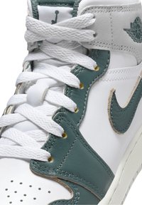 Korkeat tennarit, joissa on valkoinen nahkarunko ja teal-vihreät yksityiskohdat. Varustettu valkoisilla nauhoilla, kultaisilla nauha-aukkoilla ja rei'itetty kärki hengittävyyden lisäämiseksi.