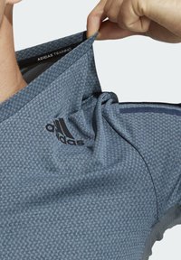 Tirando de la tela de una camiseta de entrenamiento Adidas azul por el cuello, mostrando la textura y la etiqueta interior sobre un fondo claro.