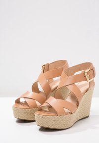 Sandalias de cuña de piel beige con tiras entrecruzadas, que cuentan con una plantilla trenzada similar a la paja y un accesorio de hebilla dorada en la correa del tobillo.