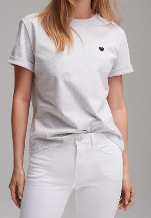 Femme portant un t-shirt gris clair à manches courtes avec un petit cœur noir sur la poitrine et un pantalon blanc ajusté sur un fond uni.