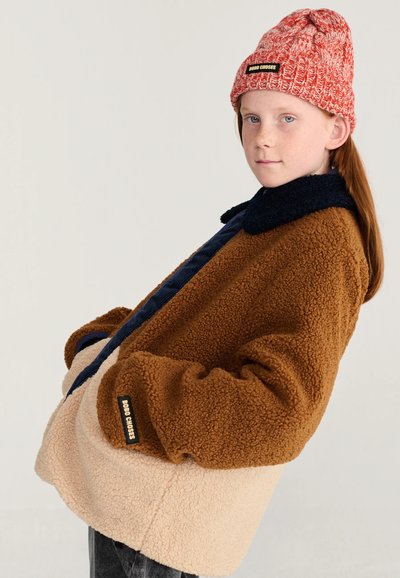 Jeune personne aux cheveux roux porte une veste polaire bicolore marron et beige et un bonnet tricoté rouge, posant les mains dans les poches sur un fond clair.