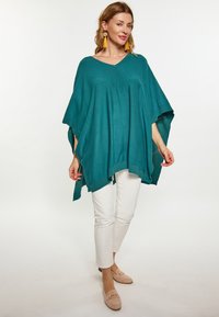 Poncho tricotat teal cu decolteu în V, având modele cu deschideri și un tiv striat. Asociat cu pantaloni albi și pantofi slip-on bej.