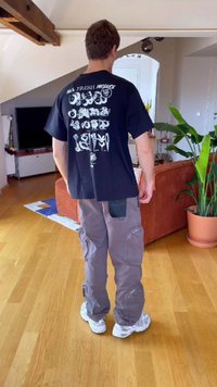 Osoba v černém tričku s bílými ilustracemi zeleniny na zádech, šedých cargo kalhotách a bílých teniskách stojí v obývacím pokoji.