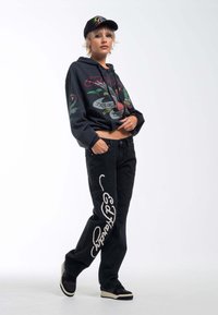 Sudadera corta negra con estampado gráfico floral y texto, combinada con jeans negros con bordado de logo blanco y zapatillas negras.