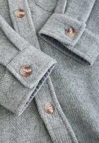 Tissu de manteau en chevron gris clair avec une texture douce, doté de boutons en or rose et de poignets pliés. Doublure subtile visible sous les poignets.