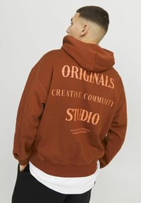 Bruine hoodie met een groot oranje grafisch tekst op de achterkant, met de woorden "ORIGINALS," "CREATIVE COMMUNITY," en "STUDIO." Zacht, textuurvezel.