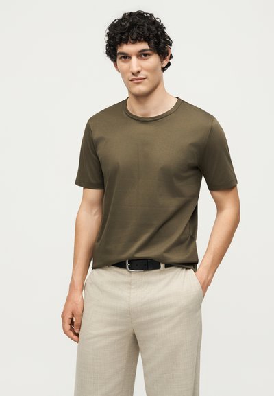 Νέος άνδρας με σγουρά σκούρα μαλλιά, που φοράει ελιάς πράσινο κοντομάνικο πουκάμισο και μπεζ παντελόνι, στέκεται με το ένα χέρι στην τσέπη, μπροστά σε απλό φόντο.