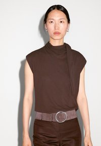 Hnědý top s krátkým rukávem a drapovaným detailem u krku. Modelka má na sobě šedý pásek s kruhovou kovovou sponou a hnědé úzké kalhoty.