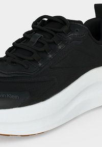 Baskets de sport noires avec semelle blanche épaisse, lacets noirs et dessus en tissu texturé, montrant les détails latéraux et du dessus.