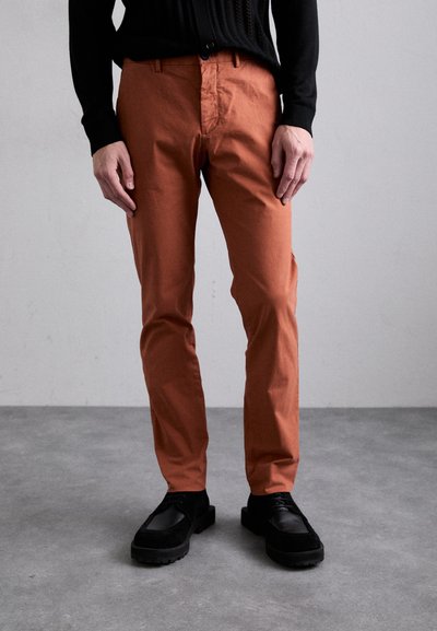 Pantalon en coton couleur rouille à coupe droite, avec une fermeture à bouton à l'avant, des poches latérales, porté avec des chaussures noires texturées.