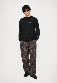Schwarzes Langarmshirt mit dem Text "NoProblemo", kombiniert mit lockeren Hosen im braun-schwarzen Camouflage-Muster und schwarzen Sneakers.