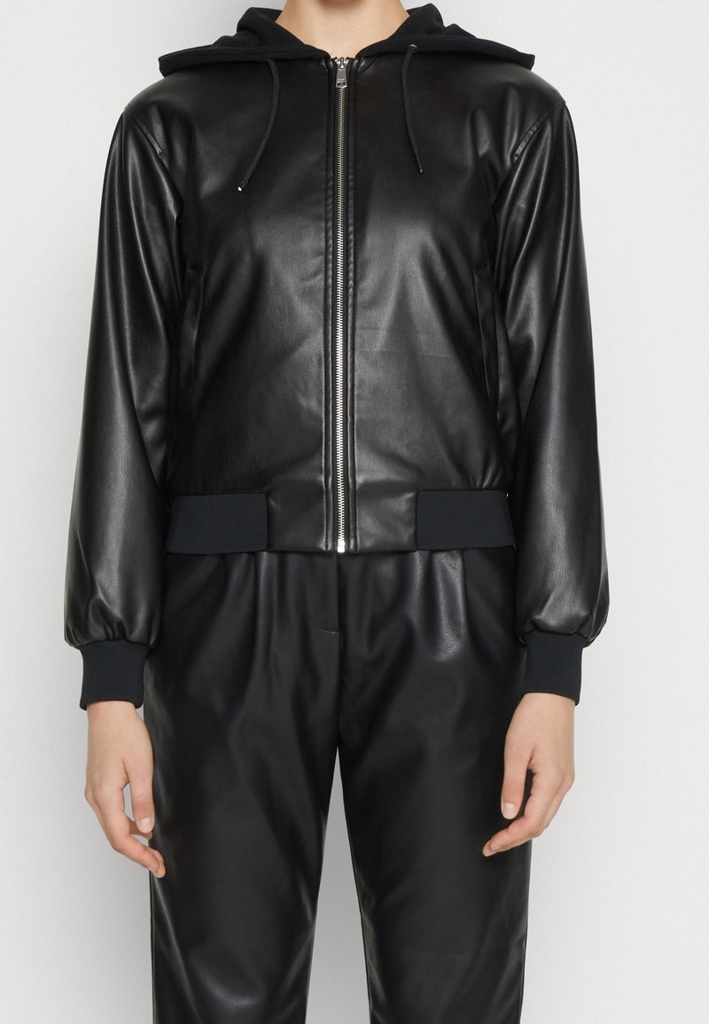 Svart fusk skinnjacka med dragkedja fram, huva och elastiska ärmslut. Slät textur med en avslappnad passform, parat med matchande svarta byxor.