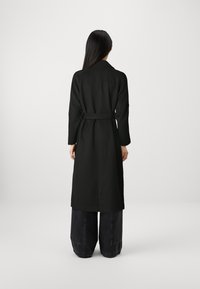 Cappotto lungo nero con cintura, colletto tradizionale e maniche ampie, abbinato a jeans larghi scuri. Texture liscia, design minimalista.