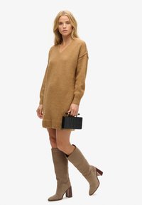 Vestido de suéter marrón de punto con escote en V y mangas largas; combinado con botas de rodilla beige y un bolso de mano negro texturizado.
