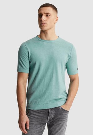 Hombre con el pelo corto que lleva una camisa de punto azul claro de manga corta y vaqueros grises, de pie con una mano en el bolsillo contra un fondo liso.