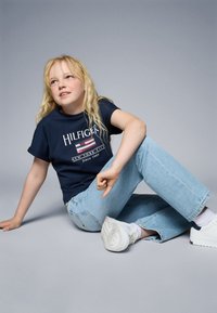 Navy cotton t-shirt with "HILFIGER" logo and American flag design, paired with light blue relaxed-fit jeans and white athletic shoes.