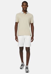 Beige pikétröja med vit krage och axelaccent, tillsammans med vita shorts. Modellen bär grå sneakers. Mjuk textur och åtsittande design.