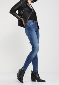 Chaqueta de cuero negra de motociclista con tachuelas plateadas, combinada con jeans ajustados azules y botas de tobillo negras con punta, sobre un fondo neutro.