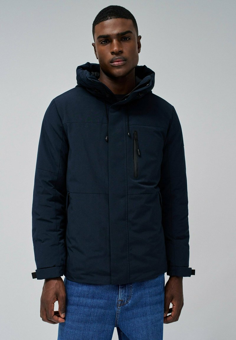 Veste isolée bleu marine avec capuche, poche zippée à l'avant et poignets ajustables. Tissu lisse avec des coutures minimales et une coupe ajustée.