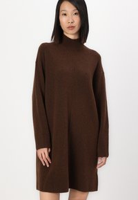 Brun strikket kjole med høy hals, løs passform og lange ermer. Stoffet har en myk tekstur og subtile variasjoner i farge.