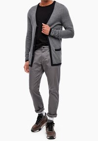 Grå stickad cardigan med svart kant, en ficka, bärs över en svart t-shirt. Kombineras med ljusgrå byxor och bruna sneakers.