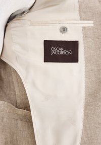 Beżowa tkanina wewnętrzna odzieży z brązową metką z logo "Oscar Jacobson" oraz srebrnym guzikiem po boku.
