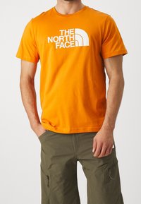 Orange bomulds t-shirt med et hvidt grafisk logo af "THE NORTH FACE" på forsiden, rund crew neck og korte ærmer.