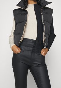 Kvinna iförd en svart pufferväst över en svart och beige ribbad långärmad topp, kombinerat med högmidjade svarta byxor med knappar, händerna i fickorna.