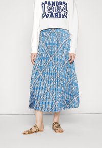 Nainen, jolla on yllään valkoinen college-paita, jossa lukee "Sandro 1984 Pariisi", ja sininen laskostettu hame, jossa on valkoisia paisleymalleja sekä vaaleanruskeat sandaalit.