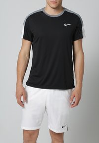 Sort atletik T-shirt med korte grå skuldre, lavet af letvægtsstof. Kombineret med hvide shorts, der har et lille logo på begge dele.