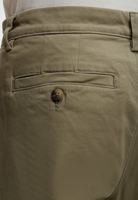 Gros plan sur la poche d'un pantalon kaki avec un rabat boutonné et des passants de ceinture visibles, porté avec une chemise côtelée beige clair.