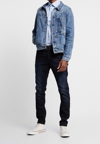 Ljus tvättad denimjacka, blåskjorta, mörka jeans och vita högsneakers. Jackan har synlig sömnad och knapprar.