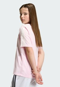 Jong meisje met lang, steil haar draagt een roze Adidas-shirt en witte korte broek, poseert met handen in elkaar gevouwen achter haar rug.