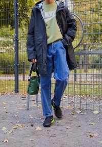 Svart vattentät jacka, grön sweatshirt med vitt Nike-logotyp, blåjeans, svarta skor med neongröna accenter och en grön väska.