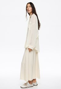 Camisa de botón ligera y transparente de color beige claro con mangas largas, superpuesta a una falda maxi ajustada de color crema. Zapatillas plateadas con diseño texturizado.
