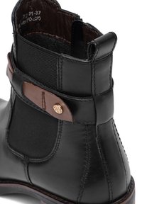 Bota de cuero negro hasta el tobillo con un panel lateral elástico texturizado, detalle de correa de cuero marrón y un adorno decorativo de botón metálico.