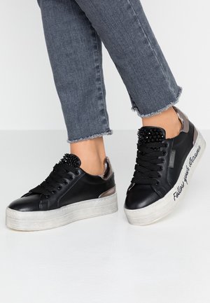 Personne portant des baskets à plateforme noires avec des languettes ornées de perles et des accents métalliques au talon, associées à un jean gris à ourlet effiloché.