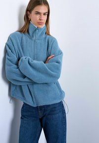 Femme appuyée contre un mur portant une veste en polaire bleue surdimensionnée avec un col haut et un jean bleu foncé, les bras croisés.