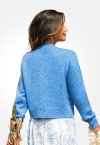 Lichtblauwe, gestructureerde gebreide trui met een ronde halslijn en lange mouwen, cropped op de taille. Een bloeiende rok en gewikkelde bloemen zijn ook zichtbaar.