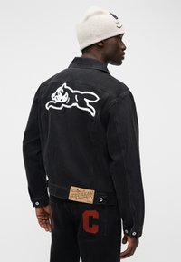 Μαύρο τζιν μπουφάν με γραφικό ενός λευκού γάτου στην πλάτη, επιβλητική ετικέτα με κεντητό σχέδιο και κόκκινη λεπτομέρεια "C" στην κάτω τσέπη.